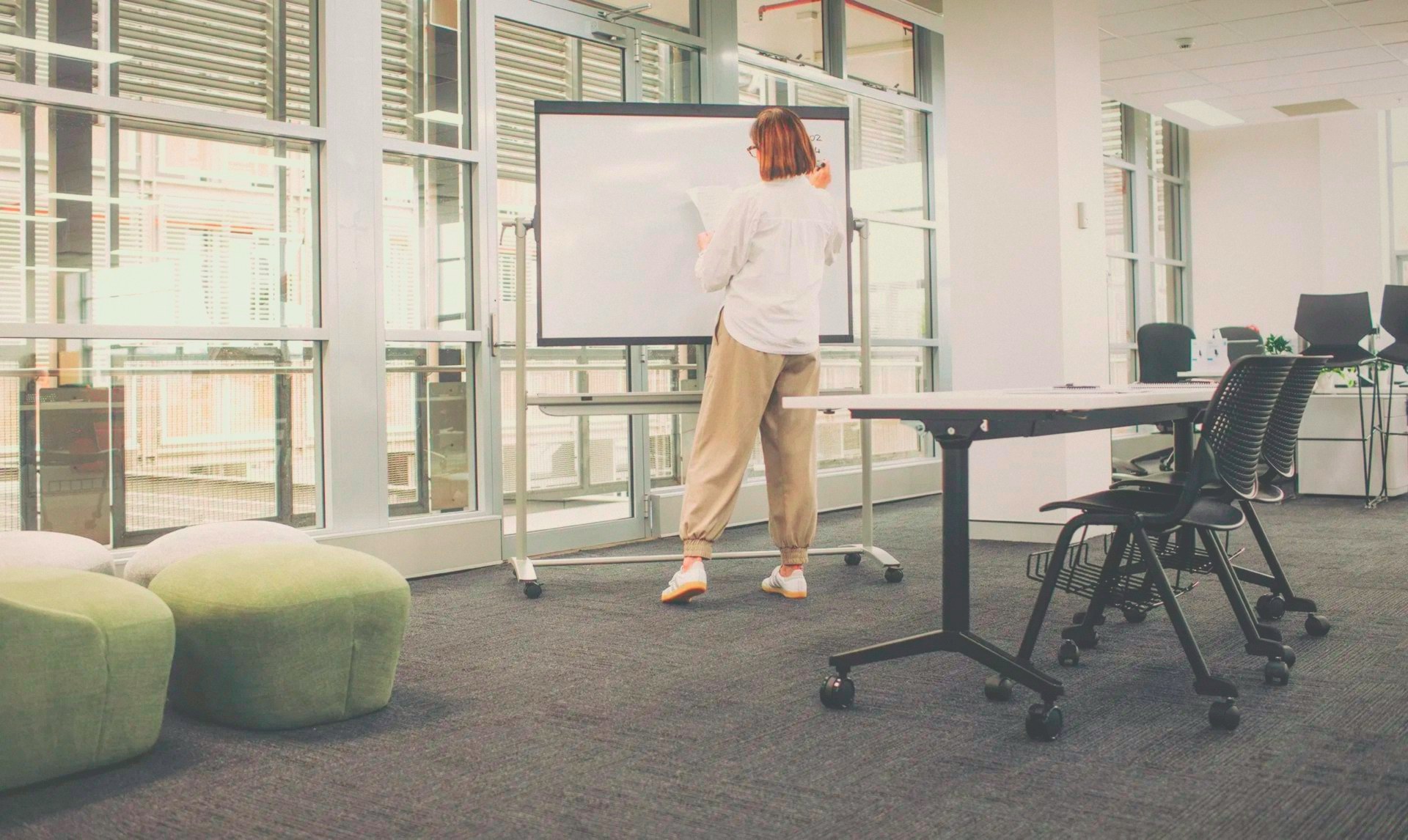 Une animatrice écrit sur un tableau blanc dans un espace de formation lumineux, avec des poufs verts et une table de travail à proximité